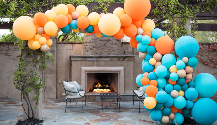 Organic Style Garland consisting of blue, orange, and yellow balloons, attached to a trellis in front of an outdoor fireplace.