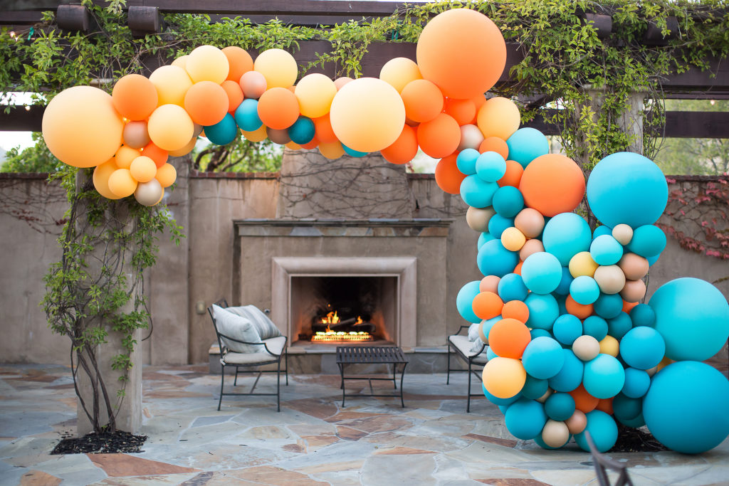 Organic Style Garland consisting of blue, orange, and yellow balloons, attached to a trellis in front of an outdoor fireplace.