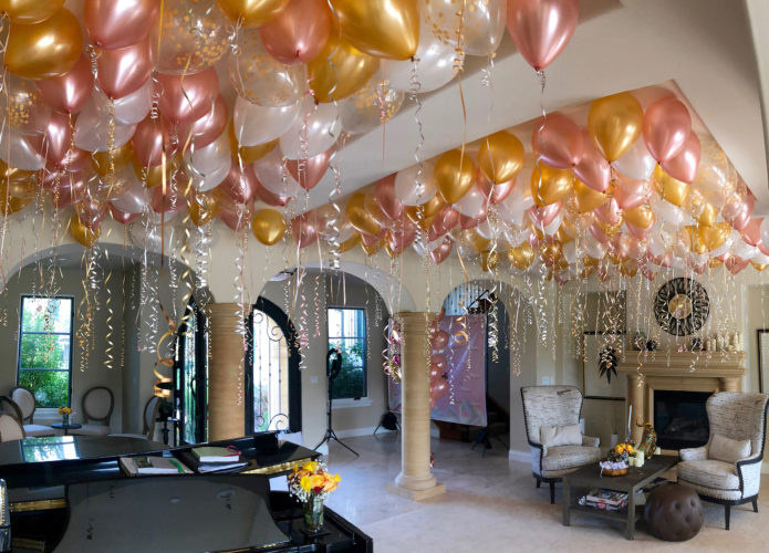 Floating rose gold, gold, pearl white, and clear balloons on matching ribbons are spread across a ceiling in a large room. The ceiling is divied into two sections by a supporting beam.