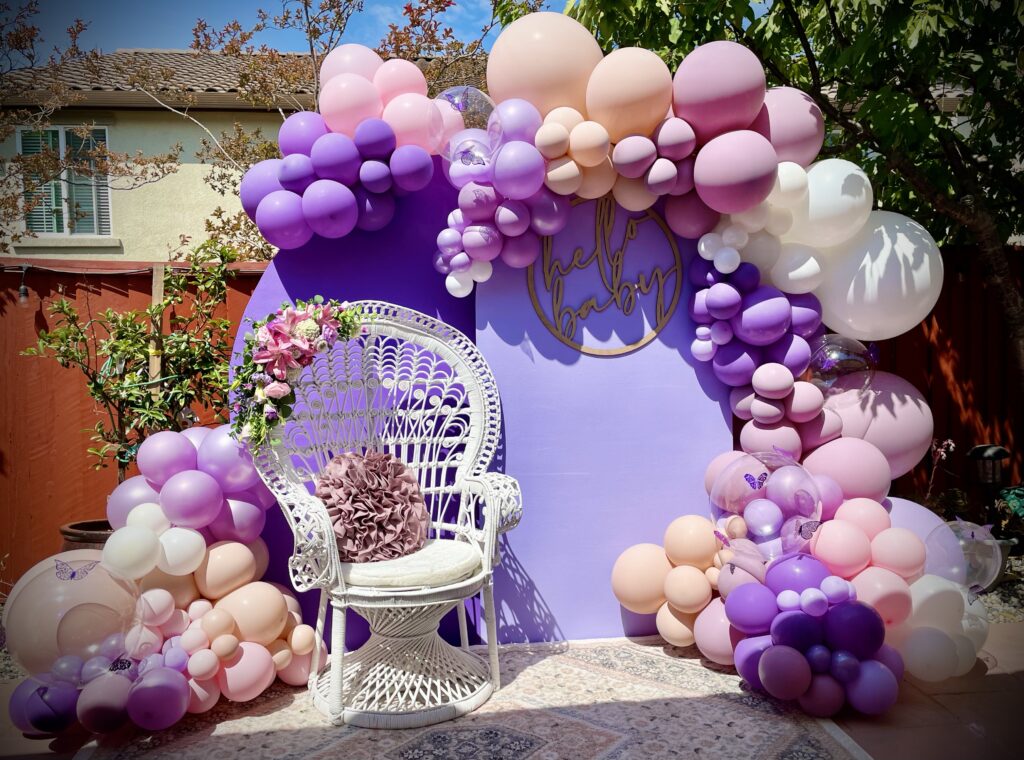 Organic Garland in shades of Purple, Pink, Blush + White including clear balloons with 3D butterfly decals, attached to purple backdrop with "Hello Baby" signage, both provided by client with an additional cluster at the bottom left corner. A white chair with florals and a purple pillow sits in front of the backdrop.