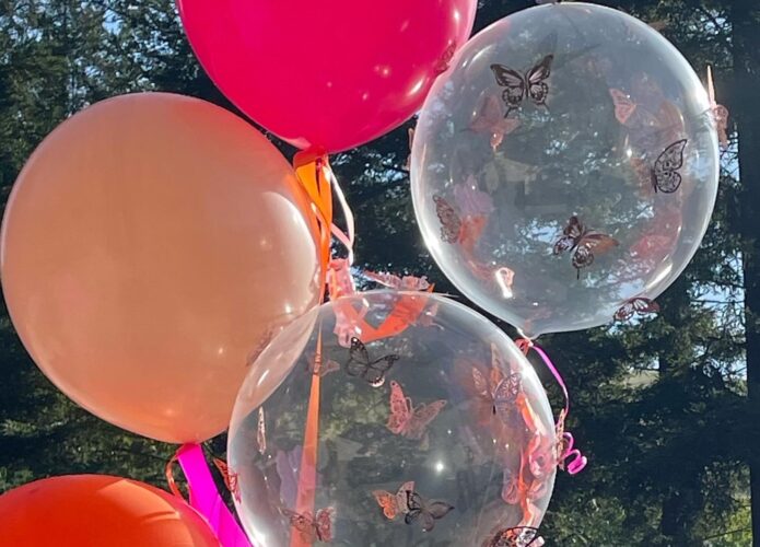 Two large clear balloons with rose gold 3D butterfly stickers, one large rose balloon, one large blush balloon, and part of a large orange balloon are grouped together outdoors. The balloons are adorned with color coordinated curled ribbons.