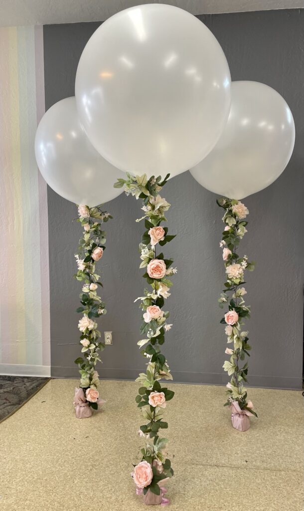 Three large white floating balloons are adorned with pink faux floral garlands and pink weights, and are placed against a grey wall.