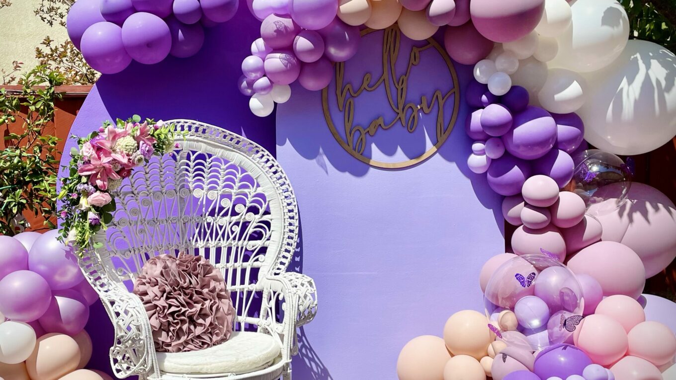 Organic Garland in shades of Purple, Pink, Blush + White including clear balloons with 3D butterfly decals, attached to purple backdrop with "Hello Baby" signage, both provided by client with an additional cluster at the bottom left corner. A white chair with florals and a purple pillow sits in front of the backdrop.