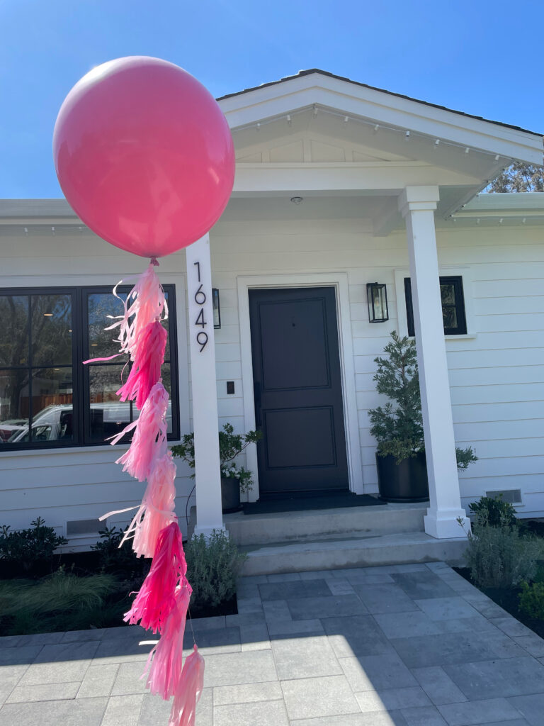 Big Pink Balloon with selection of three color pink fringy tassels hanging down attached to balloon anchored with heavy decorative weight, placed outside entrance to house to notify guests where the party is.