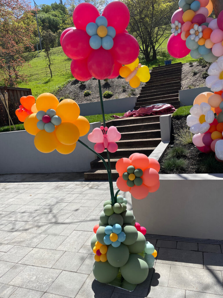 3 Blossom Balloon shaped like 5 petal flowers attached to a post in bright colors with butterfly sculptures made out of balloon hearts + green base with miniature blossom balloons