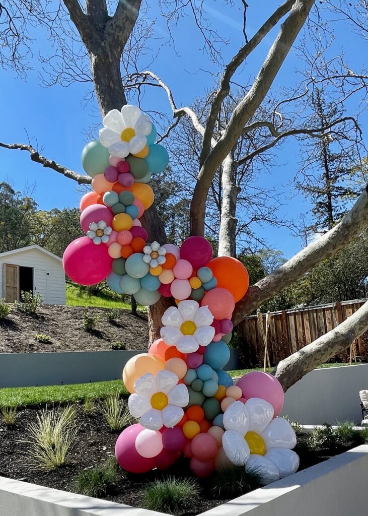 Colorful balloon garland cascading up a large tree in backyard on sunny day with blue sky in background. White Daisy Balloons stand out against the various shades of light and dark pinks with accents of orange + blush meddled with soft greens and sea glass blues.