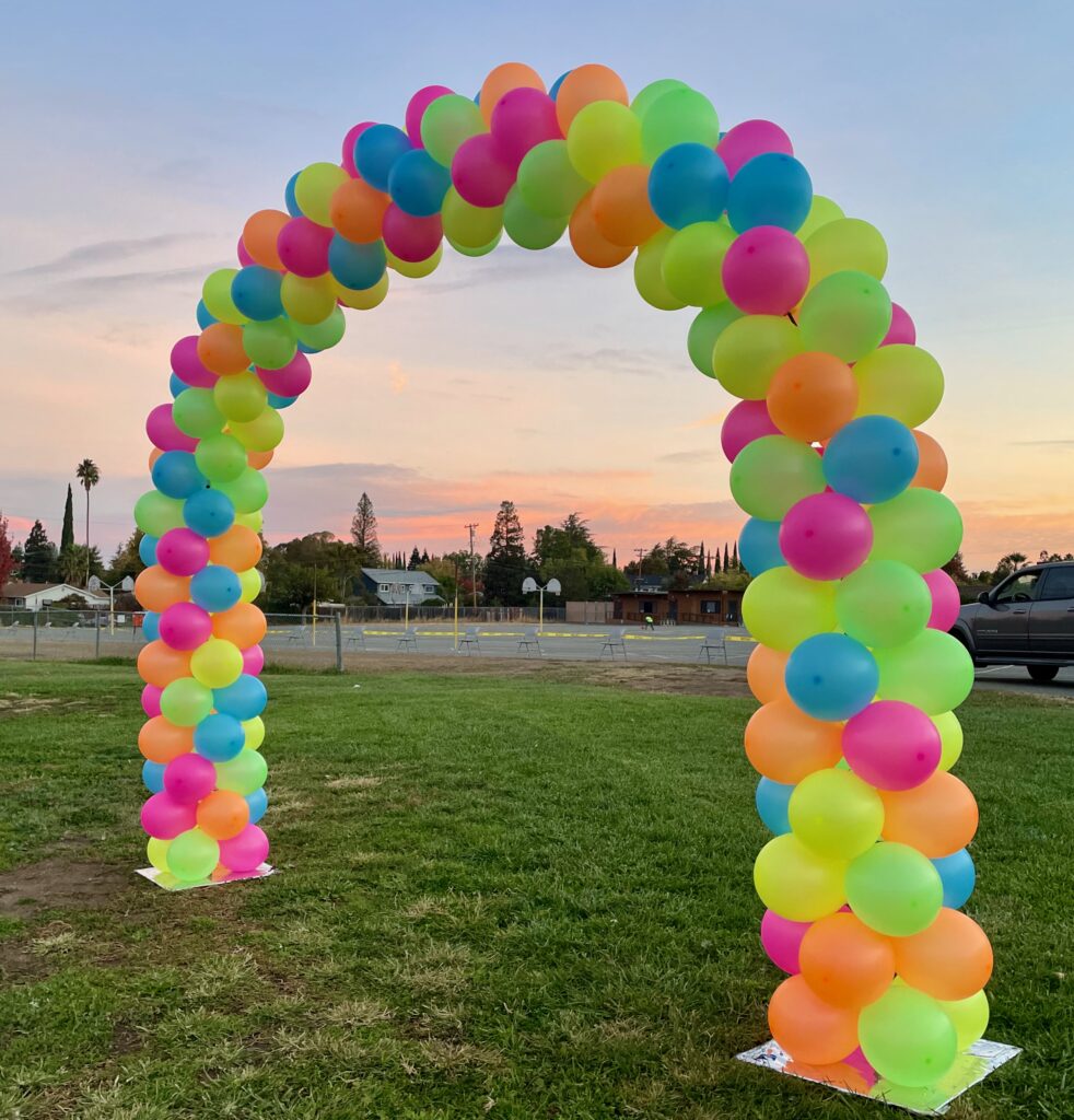 Neon balloon arch on field at sunrise. Bright orange, yellow, blue, pink, green neon colors clustered together in classic pack arch on frame. School Event for children to run through.
